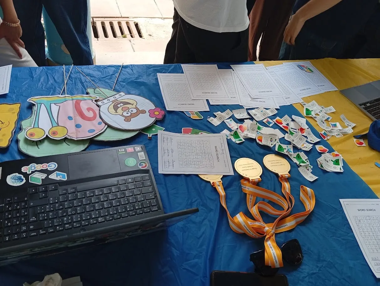 The GDG On Campus ISAMM stand: word-search sheets, Google stickers, medals on Google for Developers lanyards and a laptop covered in stickers on a blue and yellow table